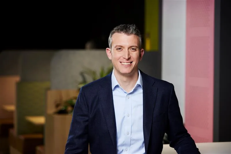 A smiling professional in a navy blazer and light blue shirt against a colorful office background with pink and yellow panels.