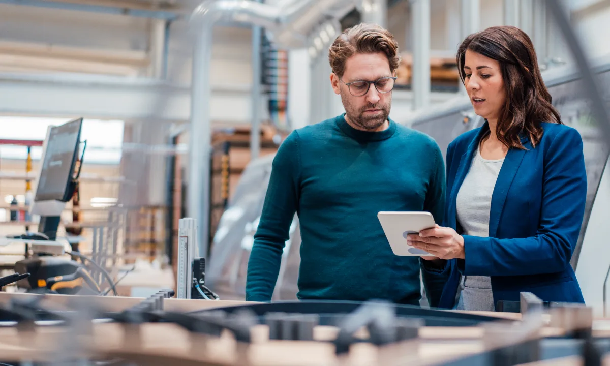 Two professionals in blue attire reviewing information on a tablet in a modern industrial workspace.