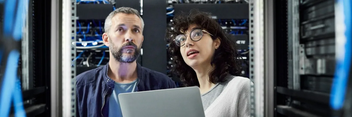 Two IT professionals looking up at server racks while one holds a laptop in a data center with blue lighting.
