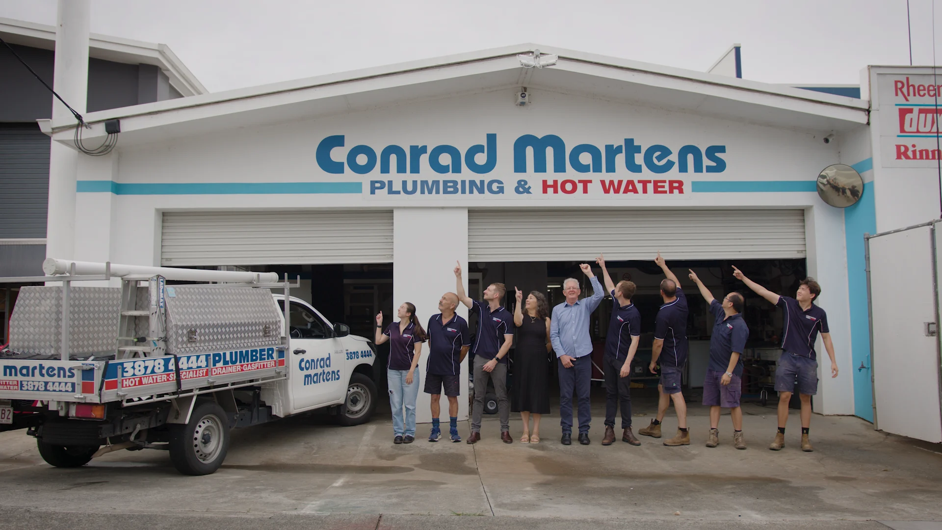 Team of plumbers in navy uniforms pointing upward outside Conrad Martens Plumbing & Hot Water storefront with company truck.