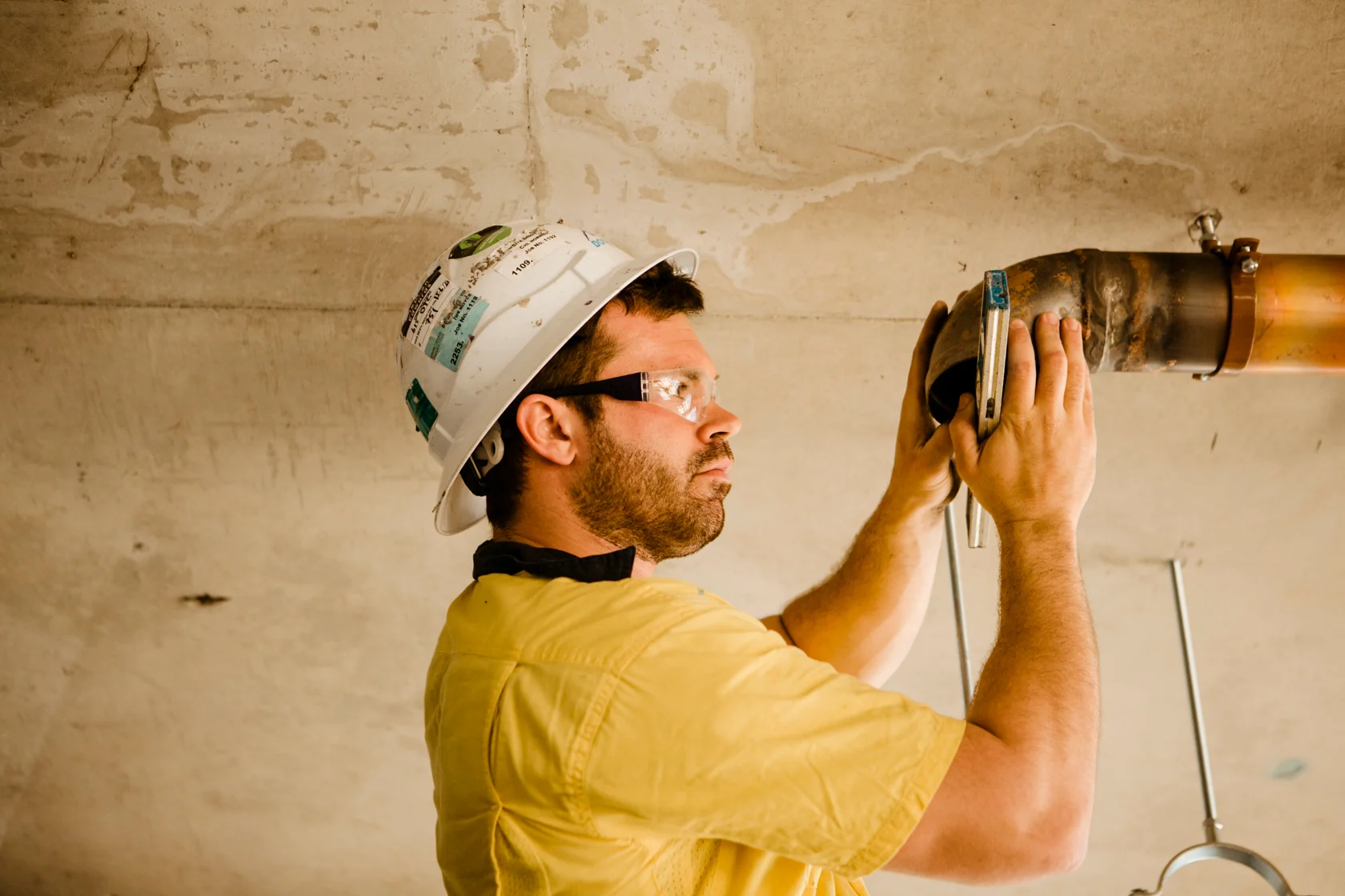 Construction worker in yellow shirt and white hard hat installing or inspecting a pipe fixture on a concrete ceiling.