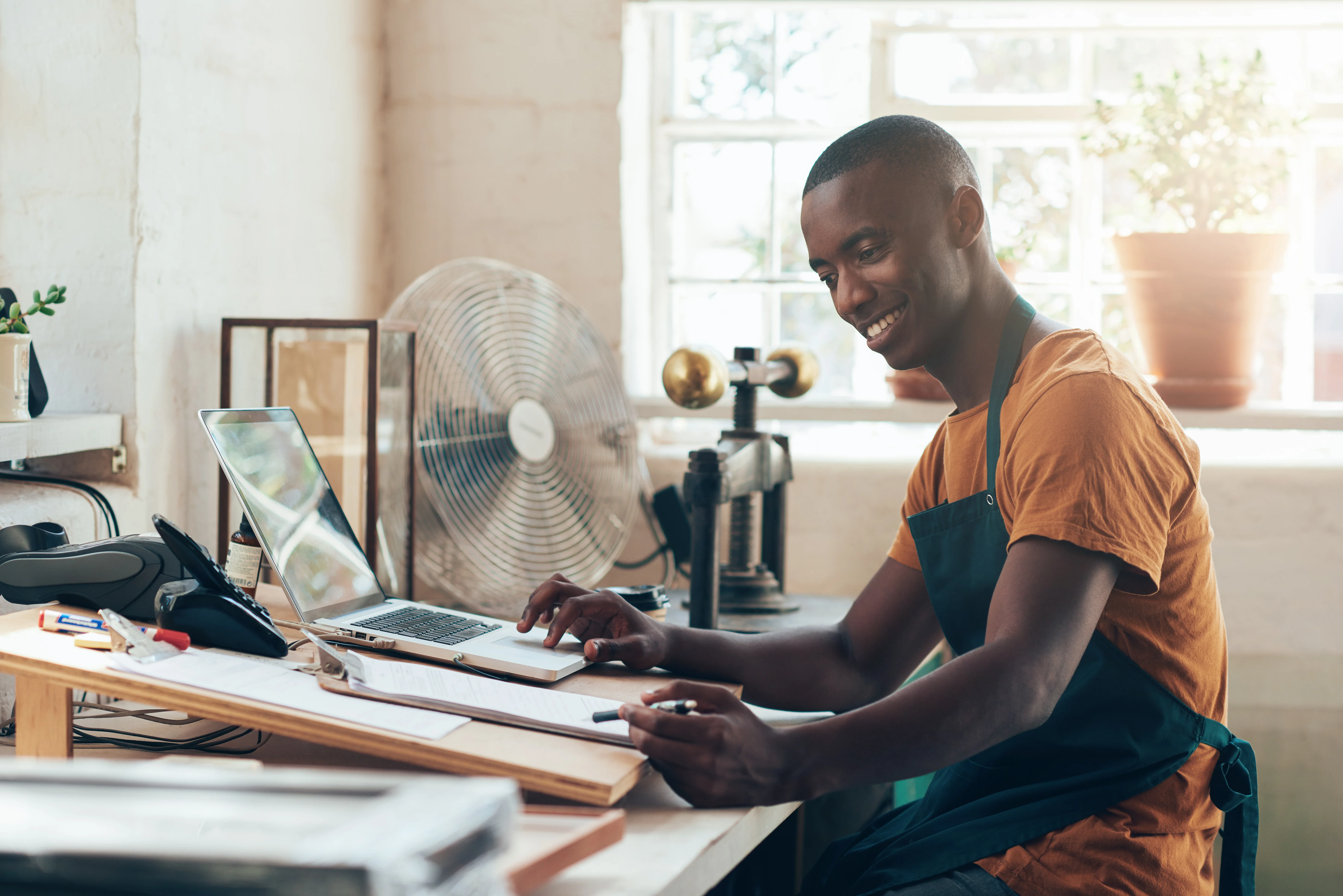 A man sitting at a work bench is using a laptop. He is smiling and wearing an apron. In the background, there is an architectural drafting board, a bench vice, a stack of picture frames, and a point of sale system on the work bench.