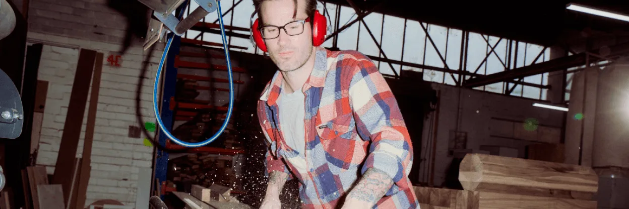 Person wearing red ear protection and plaid shirt working with wood in an industrial workshop with sawdust flying.