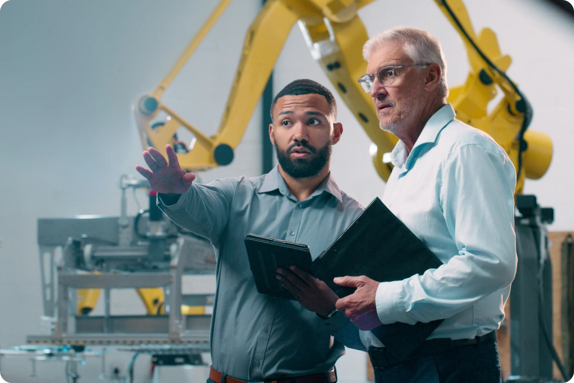 Two professionals discussing work in a manufacturing facility with yellow robotic equipment in the background.