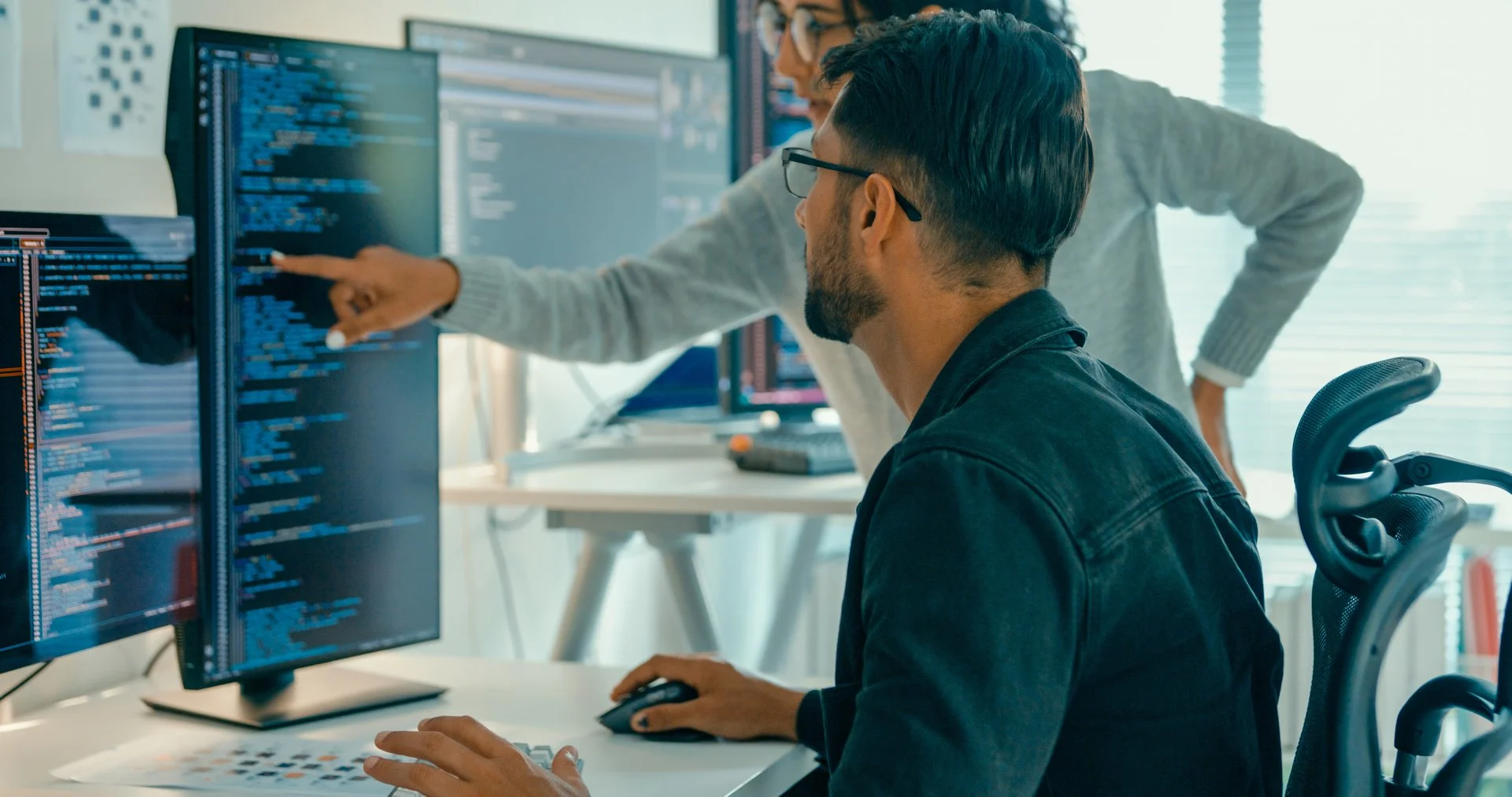 Two software developers collaborating on code, one seated at computer while colleague points at screen showing programming text.
