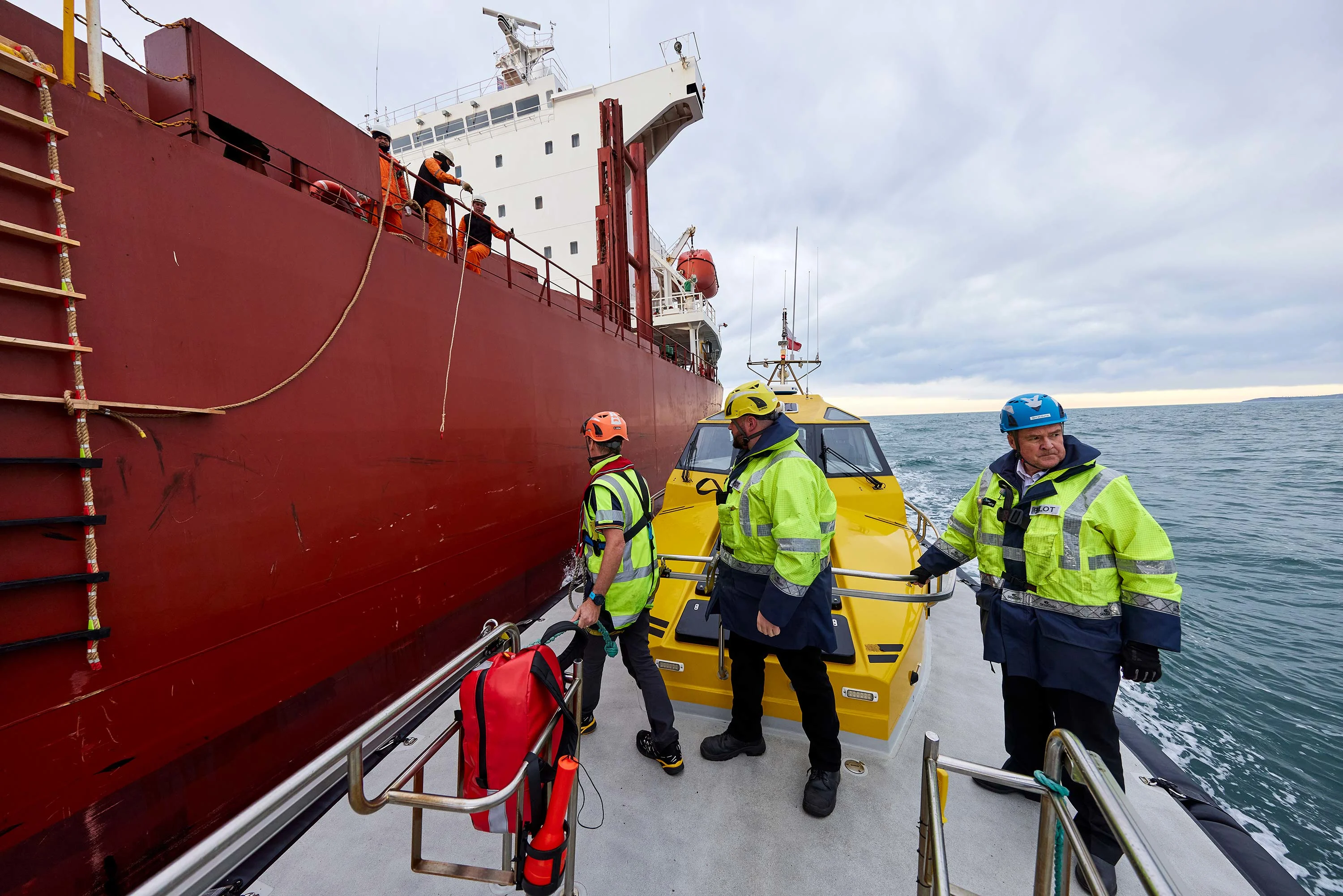 Large vessel arriving at Lyttelton Port Company. Three men in yellow jackets and hard hats in the foreground.