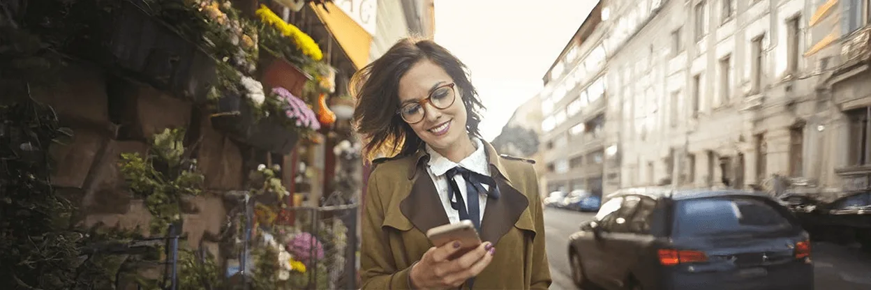 Person in tan coat checking phone while walking past flower shop on European city street with historic buildings.