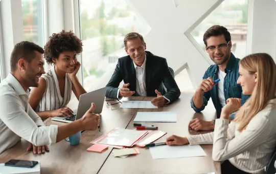 A group of 4 people sit around a meeting desk, smiling. One of them is holding a tablet computer.
