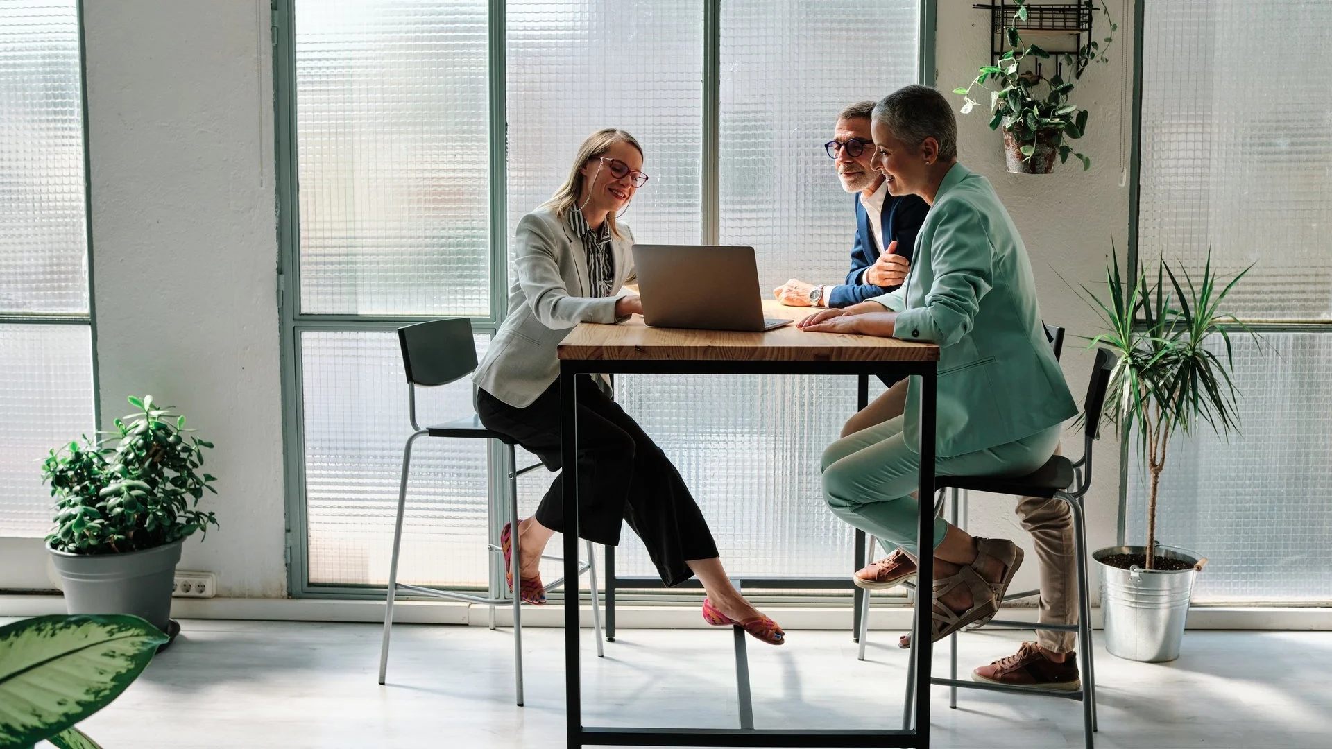 Three professionals in business attire collaborating around a laptop in a bright office space with plants.