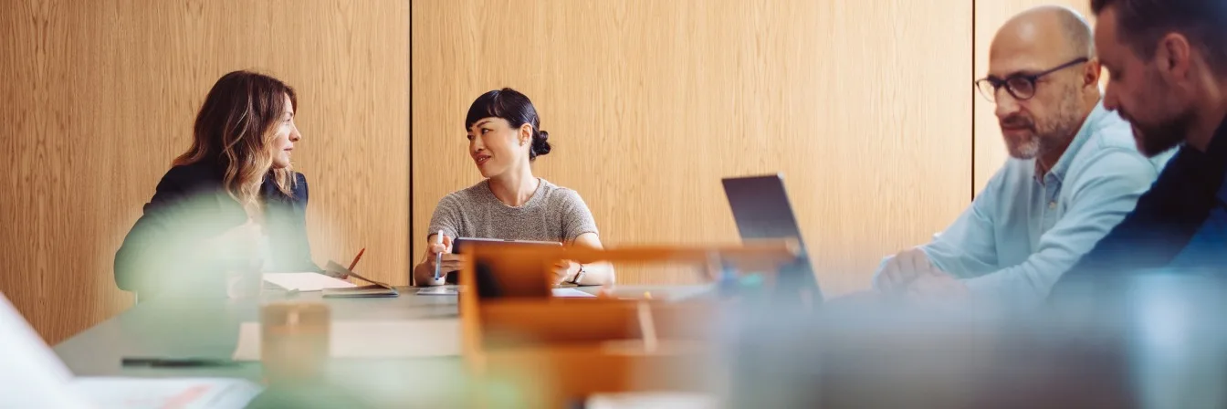 Four professionals in a modern meeting room with wooden wall panels, collaborating around a conference table.