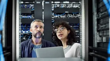 Two IT professionals examining server racks in a data center while looking at a laptop.