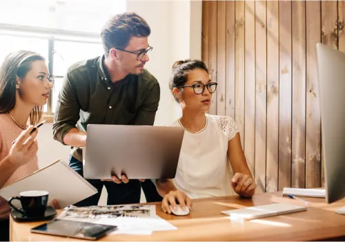 Three corporate employees gather around a desk discussing data shared on their computer screen.