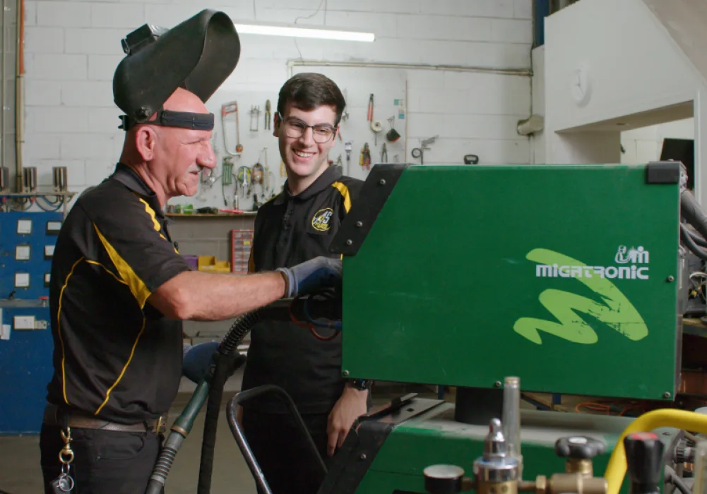 Father and son co-owners of welding supply business, A&S Welding, wearing protective gear and using a welding gun in their warehouse workshop.