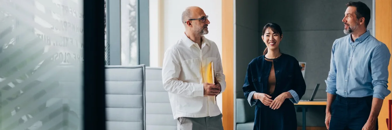 Three professionals engaged in conversation in a modern office space with large windows and minimalist furniture.