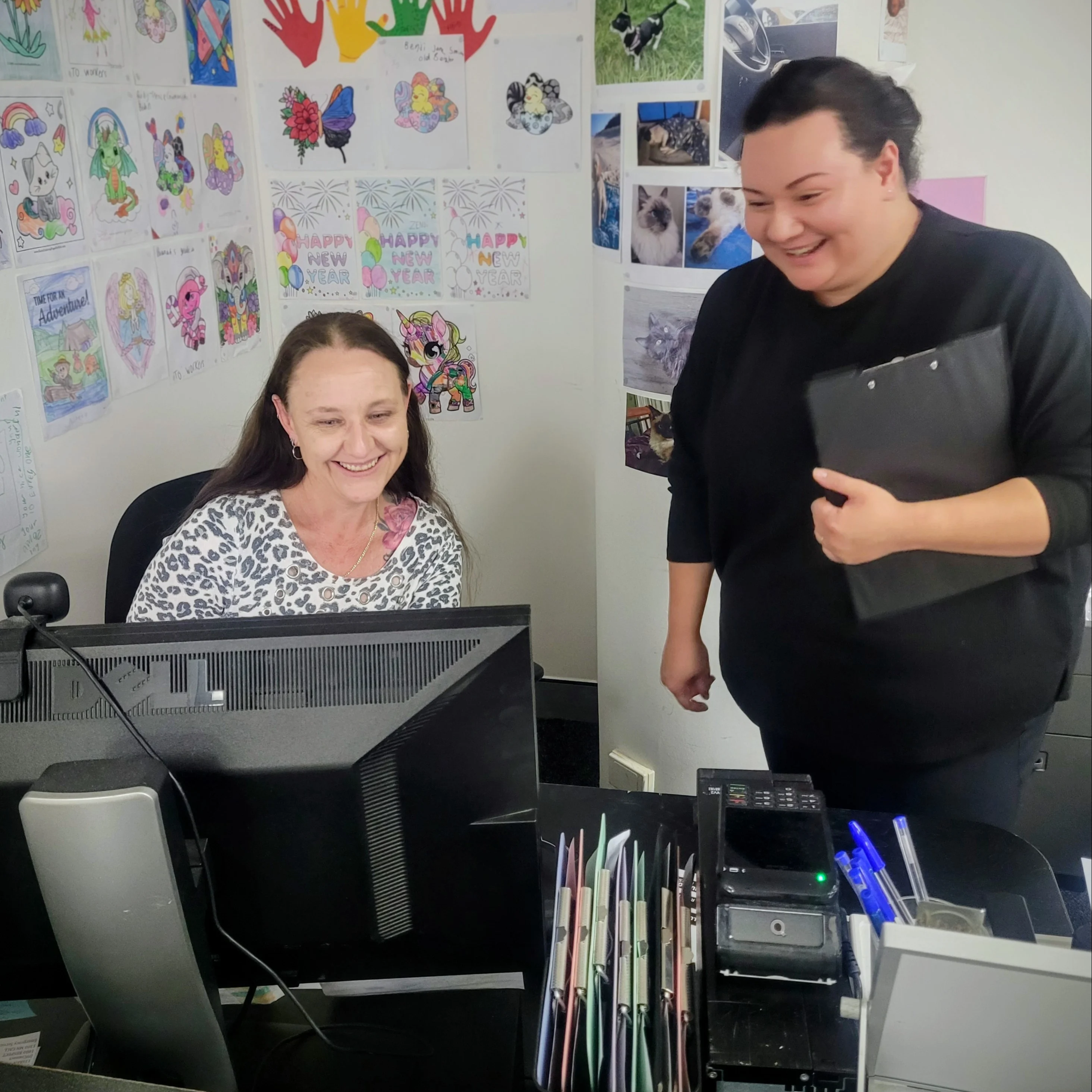 Two women smiling in an office with colorful children's artwork displayed on the wall behind them.