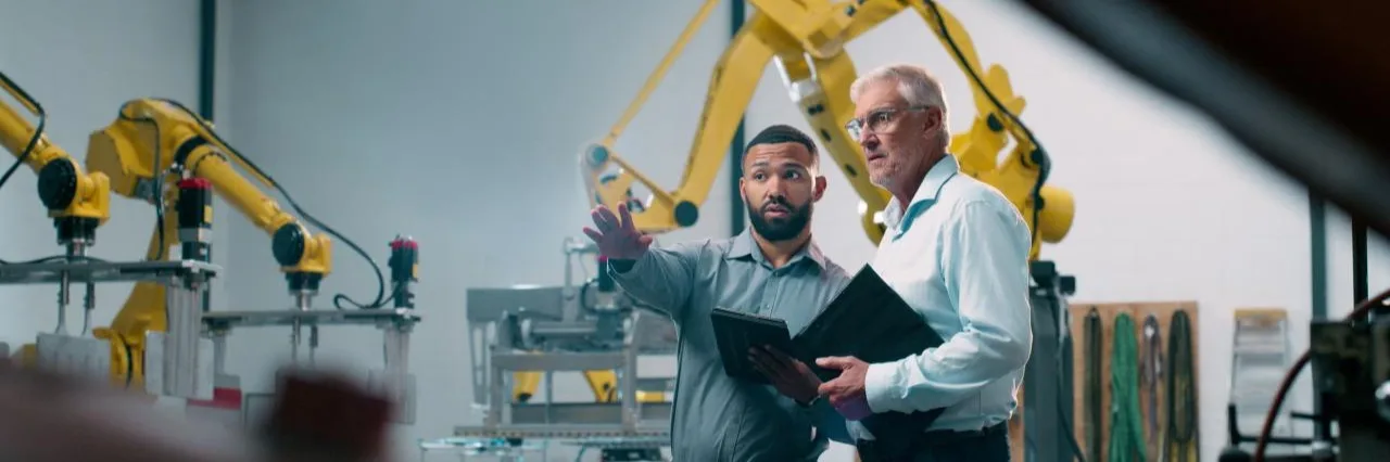 Two engineers discussing operations in a manufacturing facility with yellow robotic arms in the background.