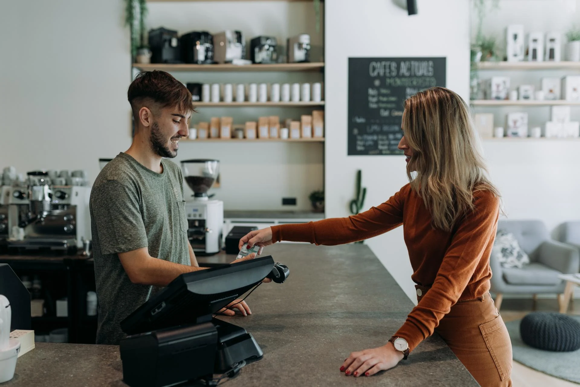Customer paying at coffee shop counter while barista in green shirt operates register, shelves of products in background.