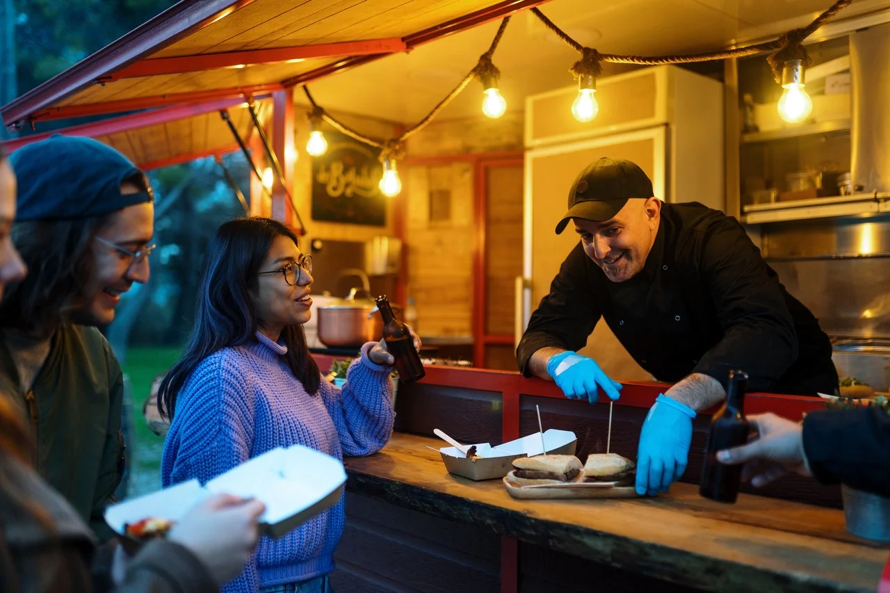 Customers ordering food at an illuminated food truck at dusk, with a chef in blue gloves serving sandwiches.