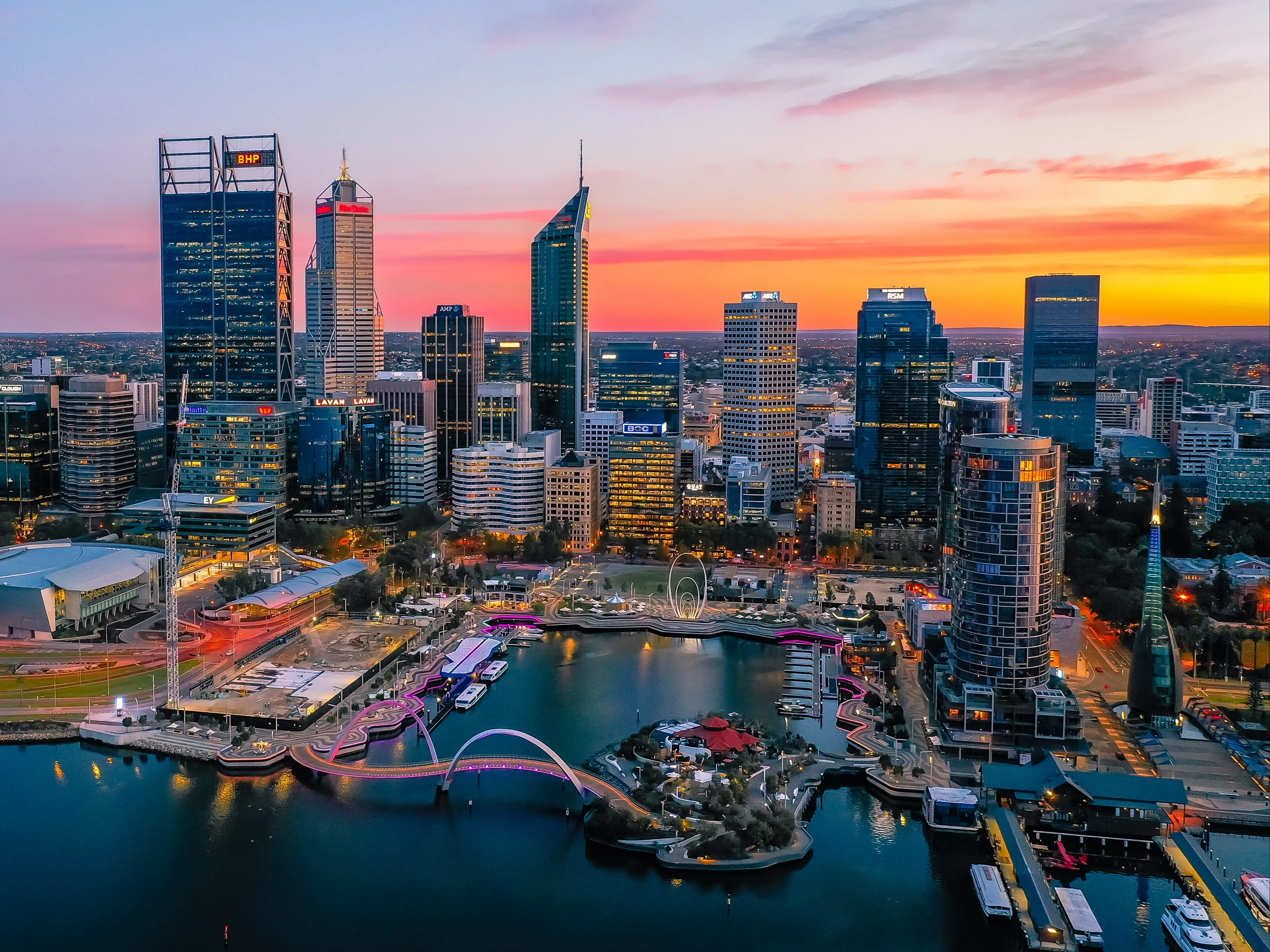 Perth skyline at sunset with illuminated skyscrapers and Elizabeth Quay waterfront featuring colorful lighting.