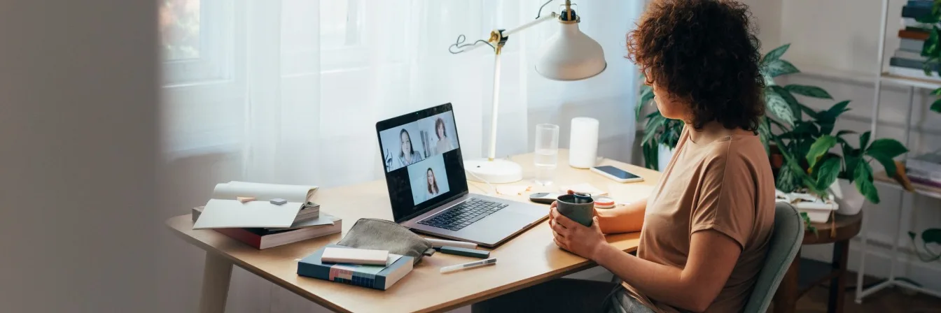 Person with curly hair on video call at home desk with laptop, books, lamp, and plants in bright workspace.