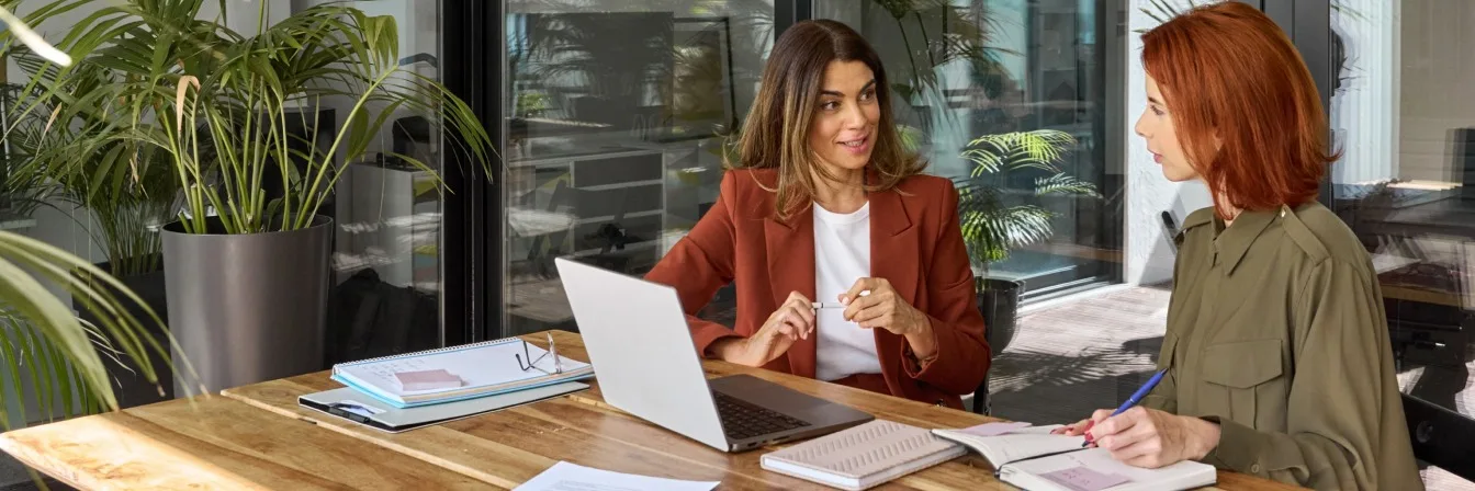 Two women in business meeting at wooden table with laptop, notebooks and plants in bright modern office space.