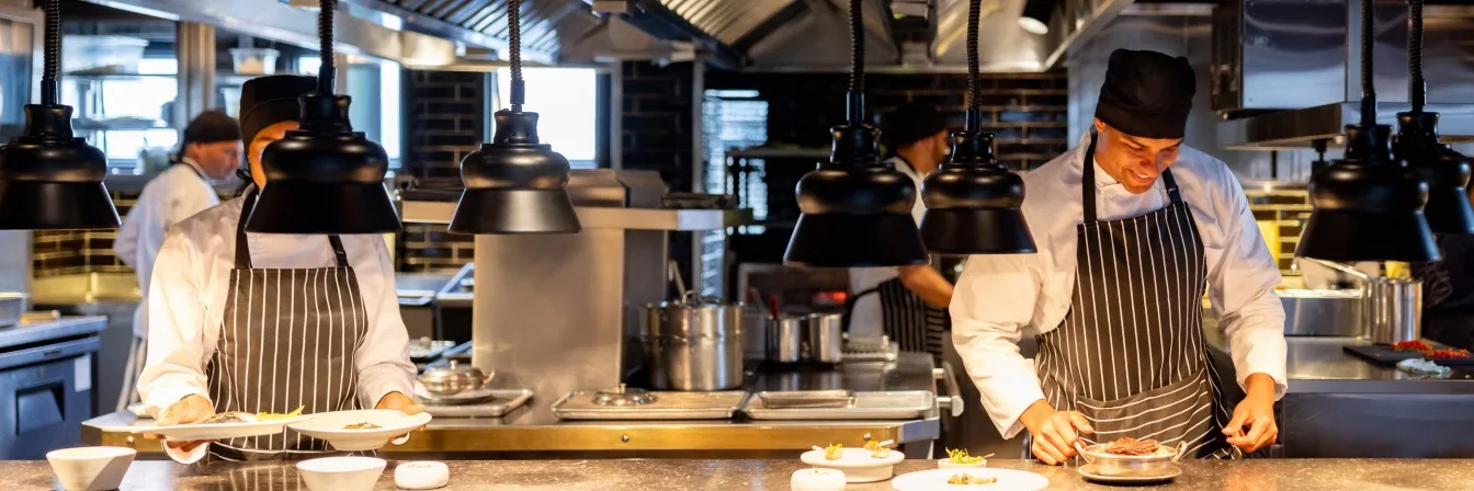 Chefs in striped aprons working in a professional kitchen with hanging black lamps and stainless steel equipment.