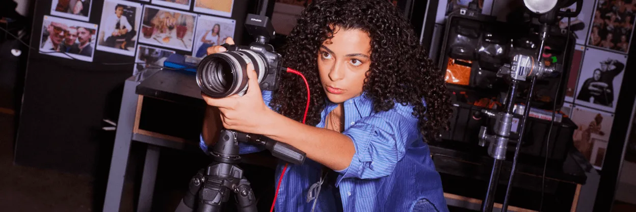 Person with curly hair operating a Canon camera on tripod in photography studio with photos displayed on wall behind.