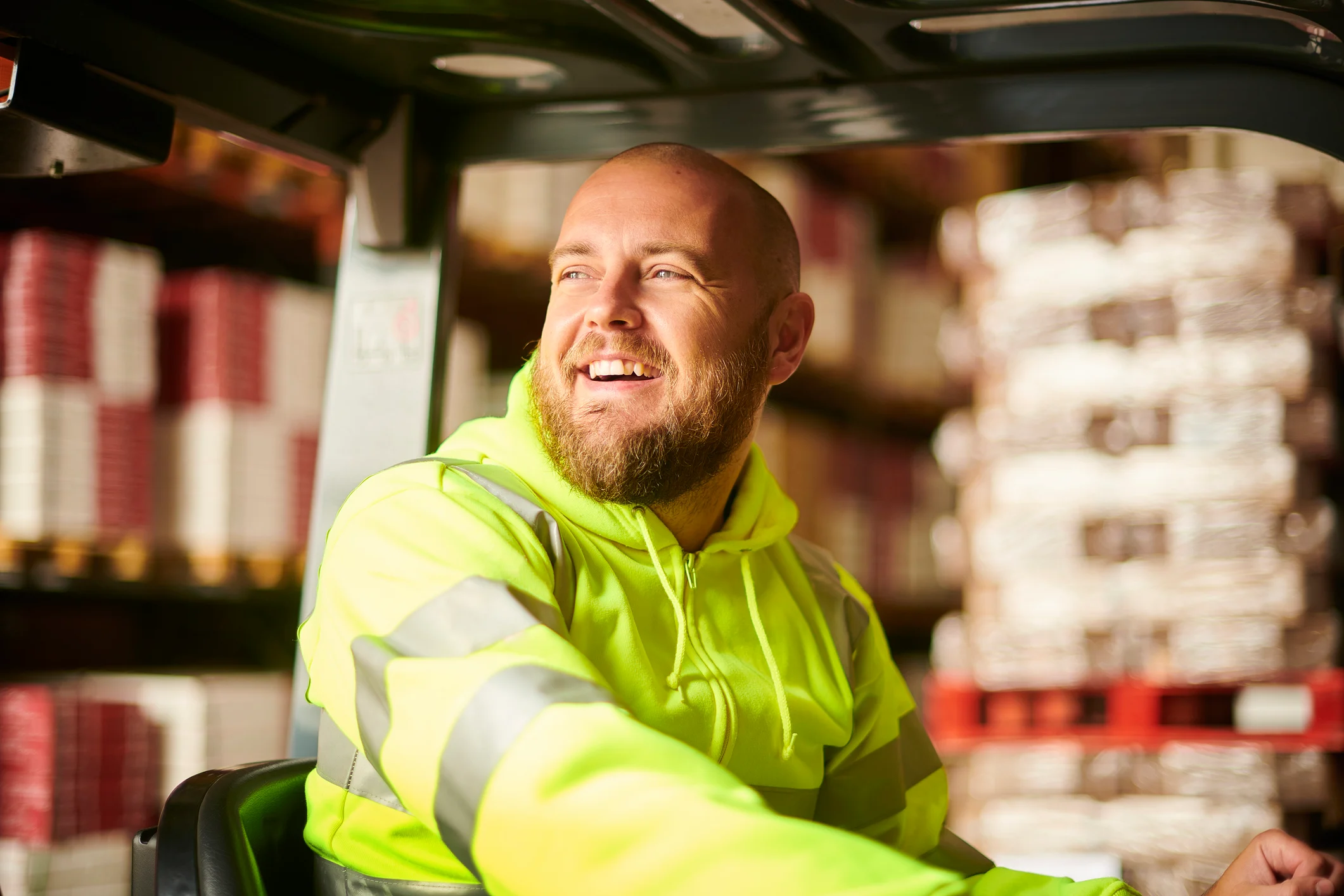 A man in high visibility clothing driving a truck past delivery palettes and smiling.