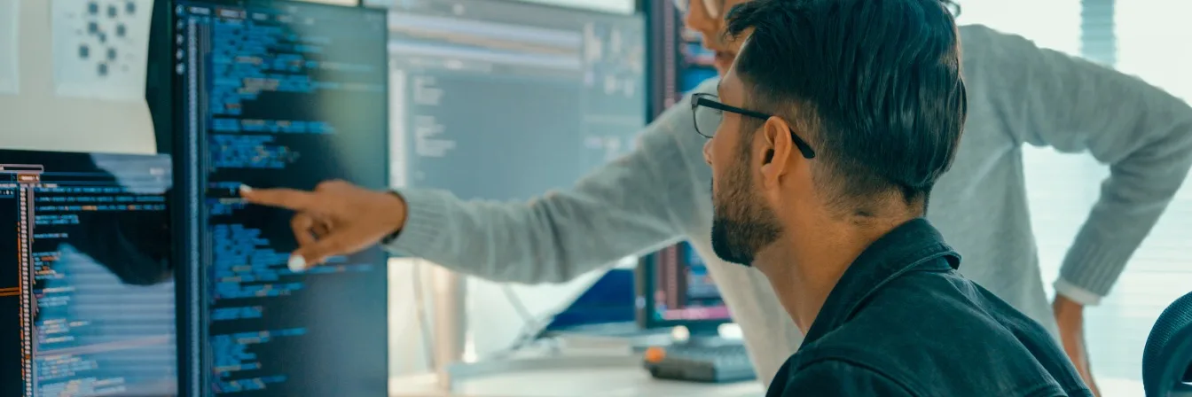 Two software developers reviewing code on multiple computer screens in a bright modern office environment.