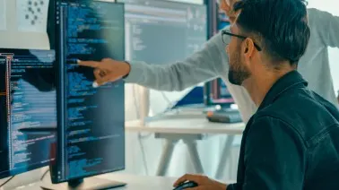 A person pointing at code on a computer screen while working at a desk with multiple monitors in an office setting.