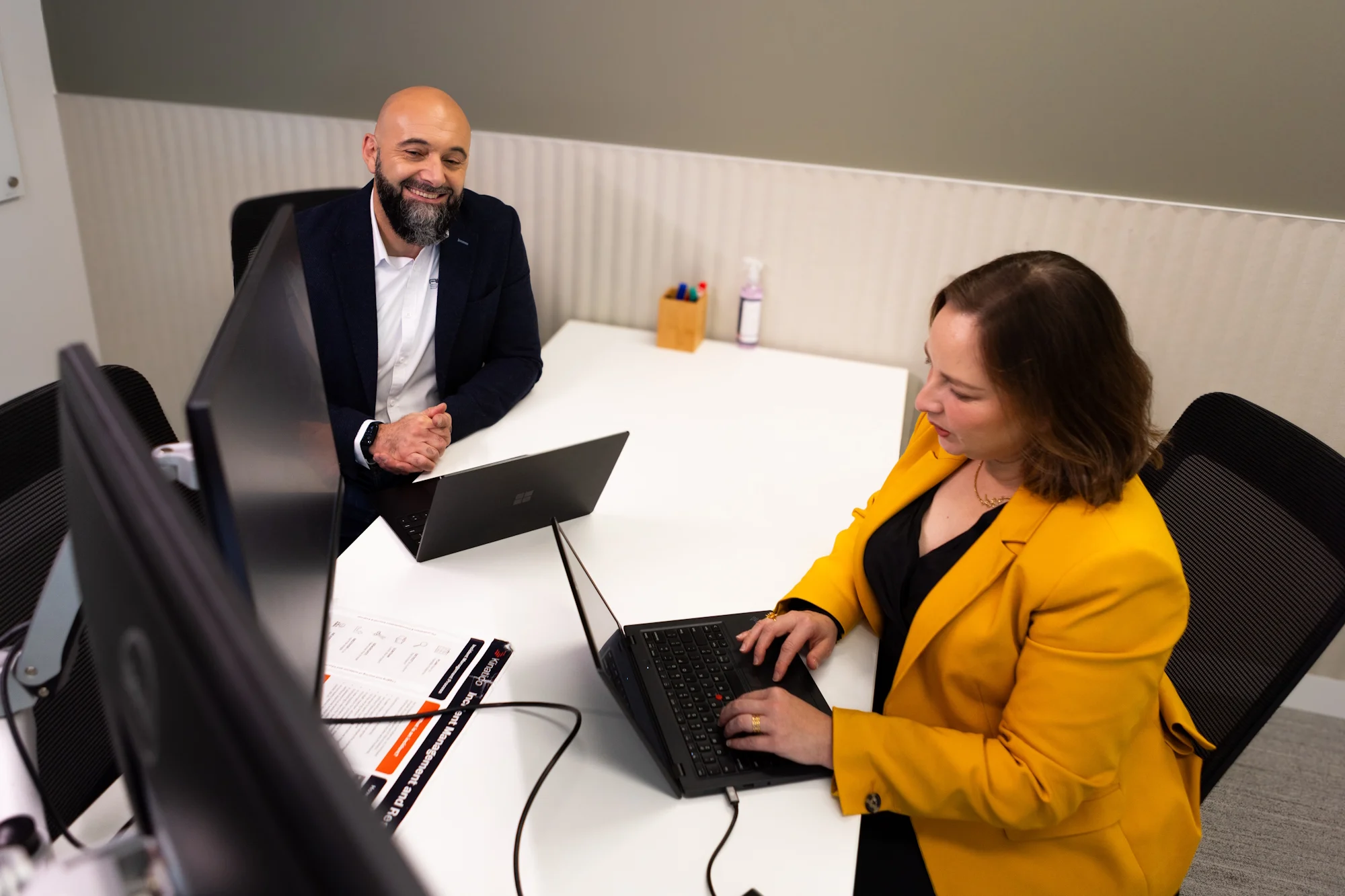 Two professionals in an office meeting, man in navy blazer smiling while woman in bright yellow jacket works on laptop.