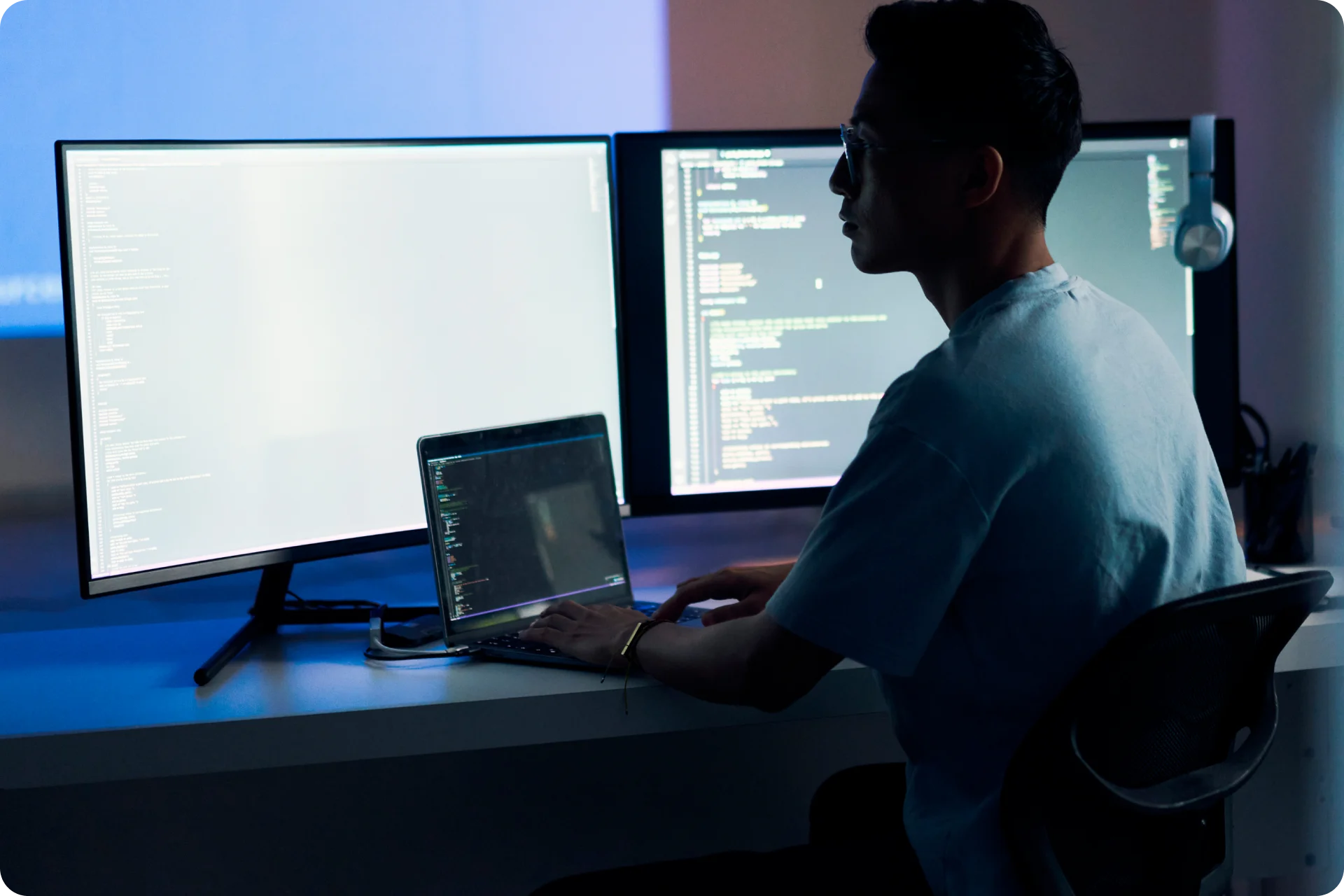 Person working at night with dual monitors and laptop displaying code in a dimly lit room with blue ambient lighting.