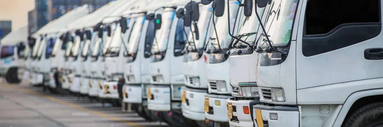 Row of white delivery trucks parked in line at a commercial depot, with cabs and side mirrors visible.
