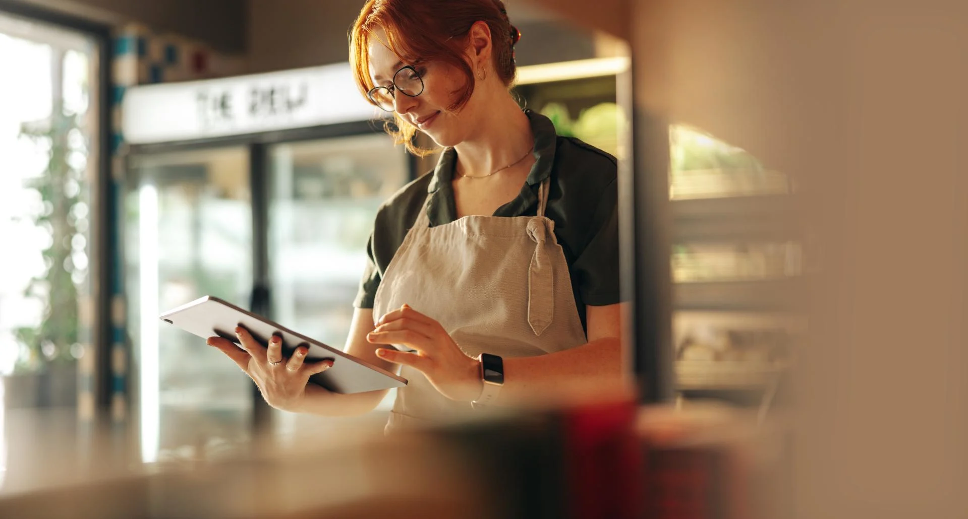 Person with red hair and glasses wearing beige apron using tablet in a café or small business setting.