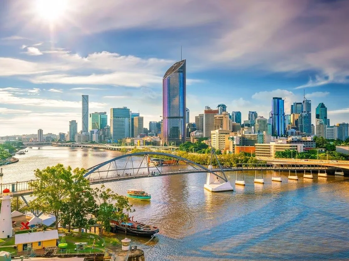 Brisbane skyline with modern skyscrapers, Brisbane River, and pedestrian bridge under bright blue sky with sunlight.