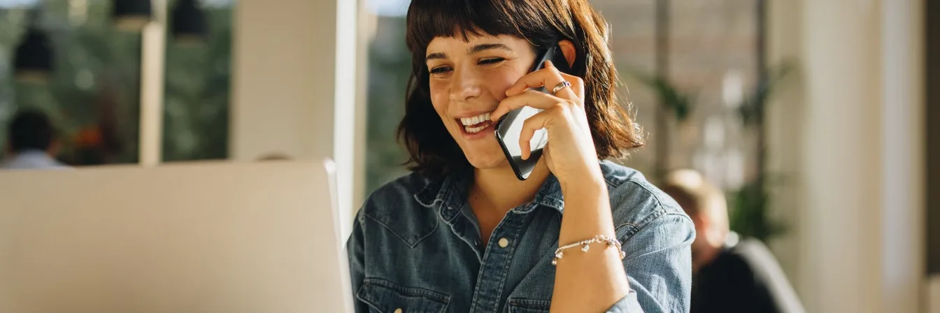 Person in denim shirt smiling while talking on phone in bright office space with coffee cup nearby.