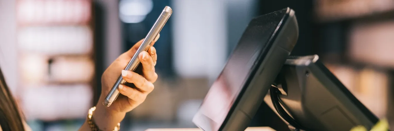 Hand holding smartphone next to a point-of-sale terminal in a retail environment, suggesting mobile payment.