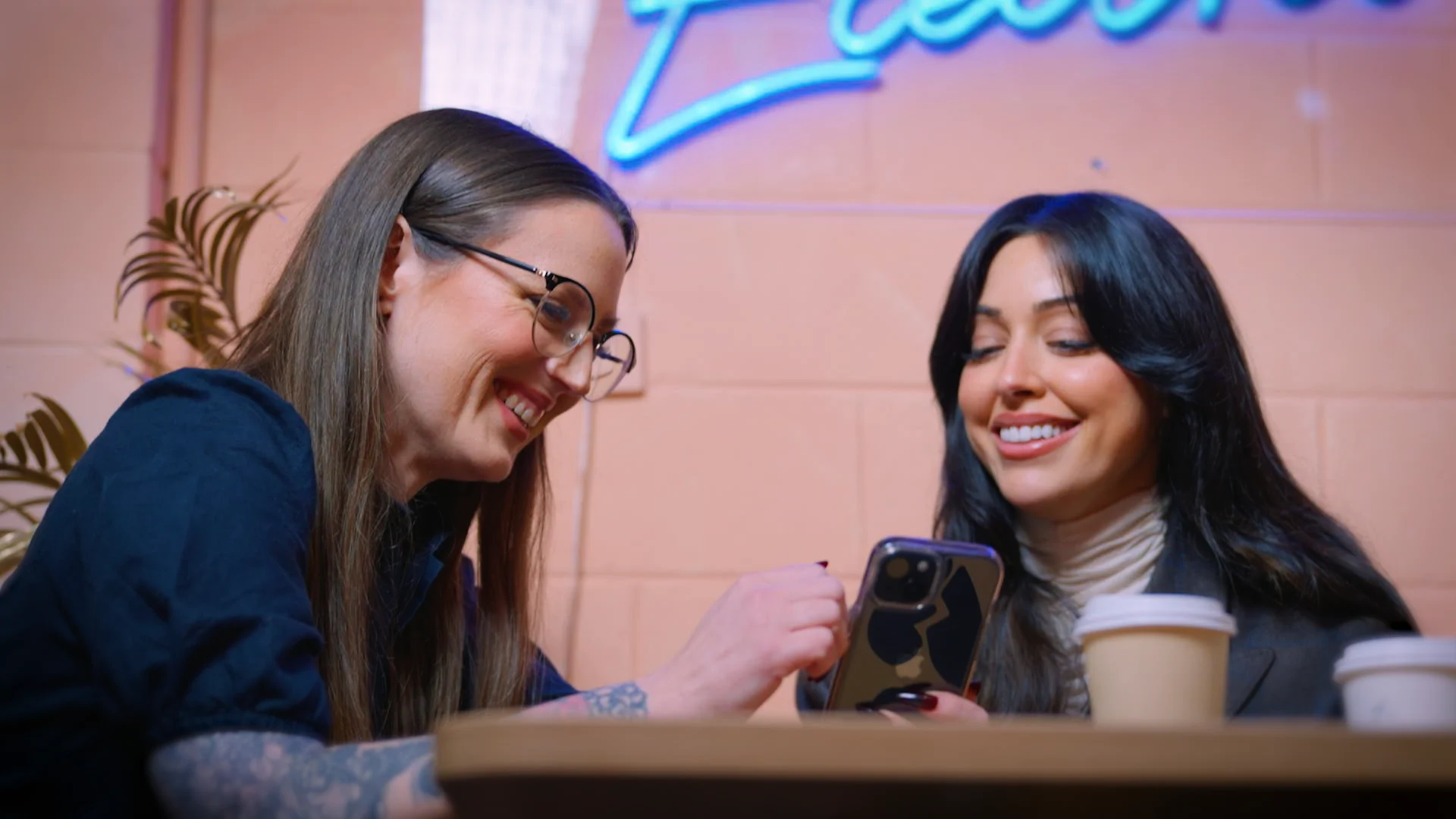 Two women smiling while looking at a smartphone together in a café with pink walls and blue neon sign.