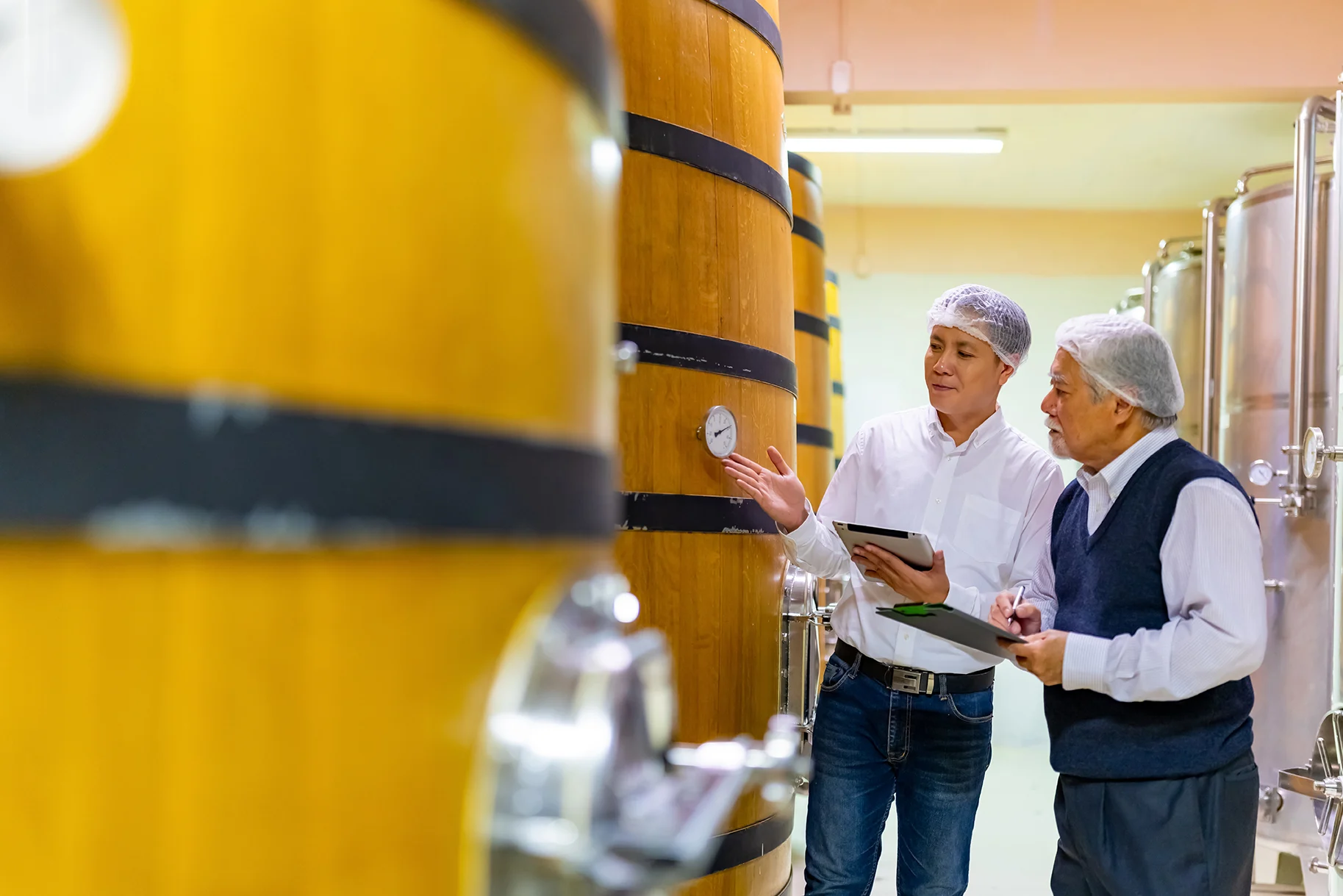 Image of two men wearing hair nets and carrying clipboards, inspecting the gauge on one of many vats of wine.