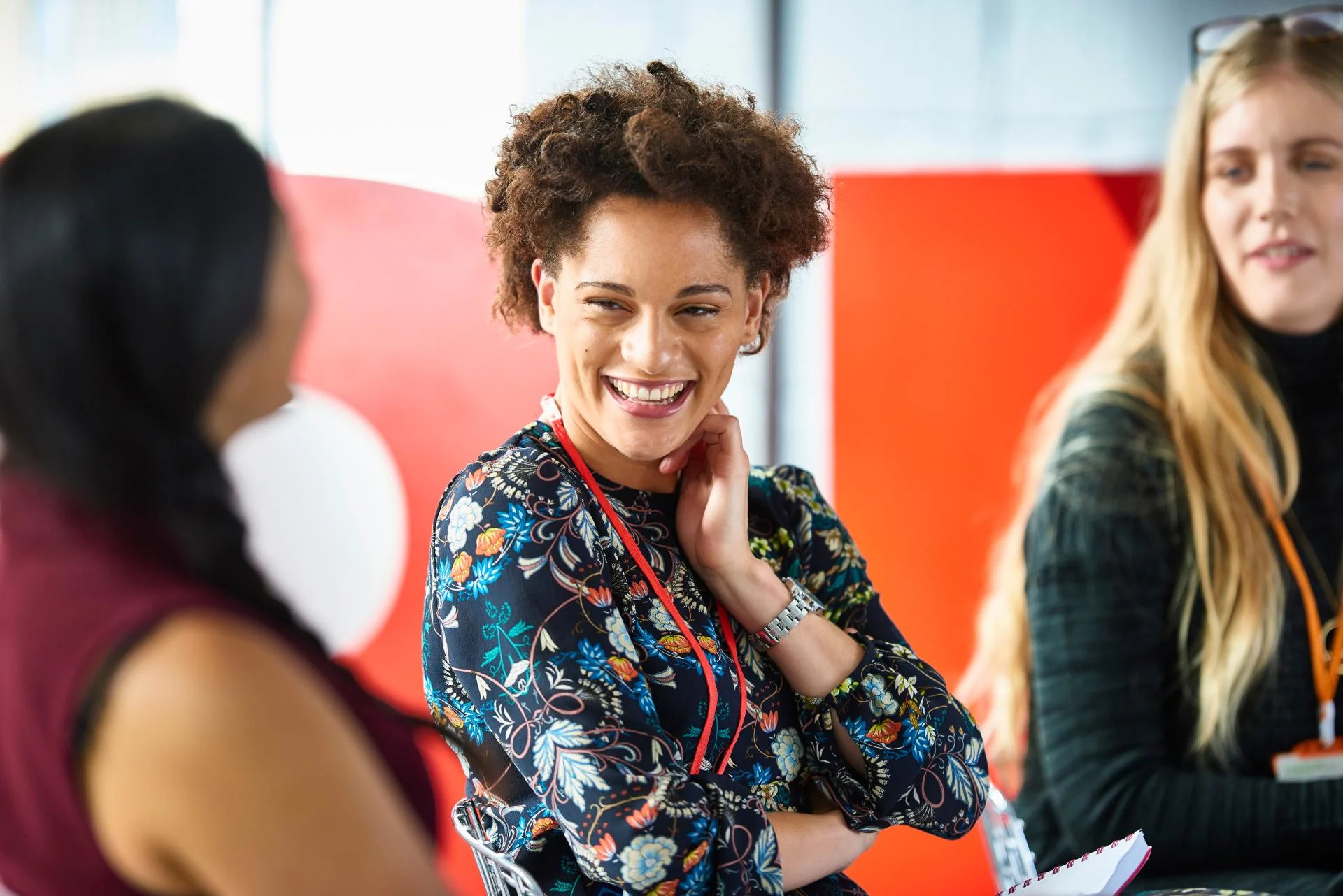 Woman with curly hair in floral blouse laughing during conversation with colleagues against red background.