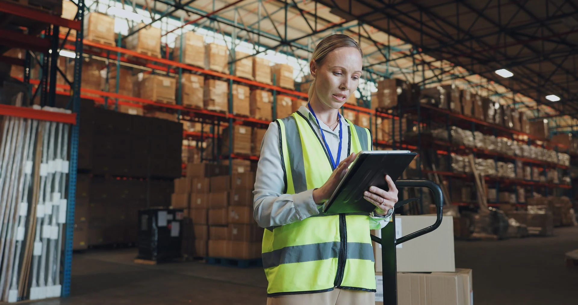 Warehouse worker in safety vest using tablet to manage inventory with stacked boxes on shelves in background.