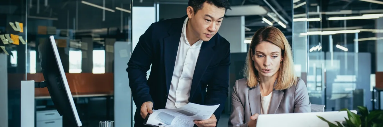 Two professionals in business attire collaborating at a desk with computer and documents in a modern office space.