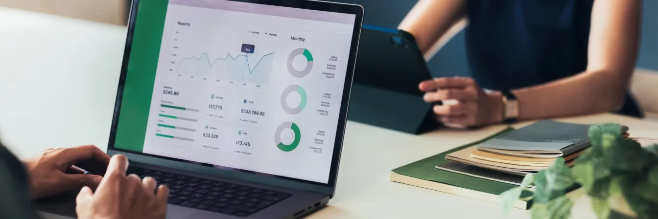 Laptop displaying financial dashboard with graphs, charts and revenue figures while someone reviews data during a meeting.