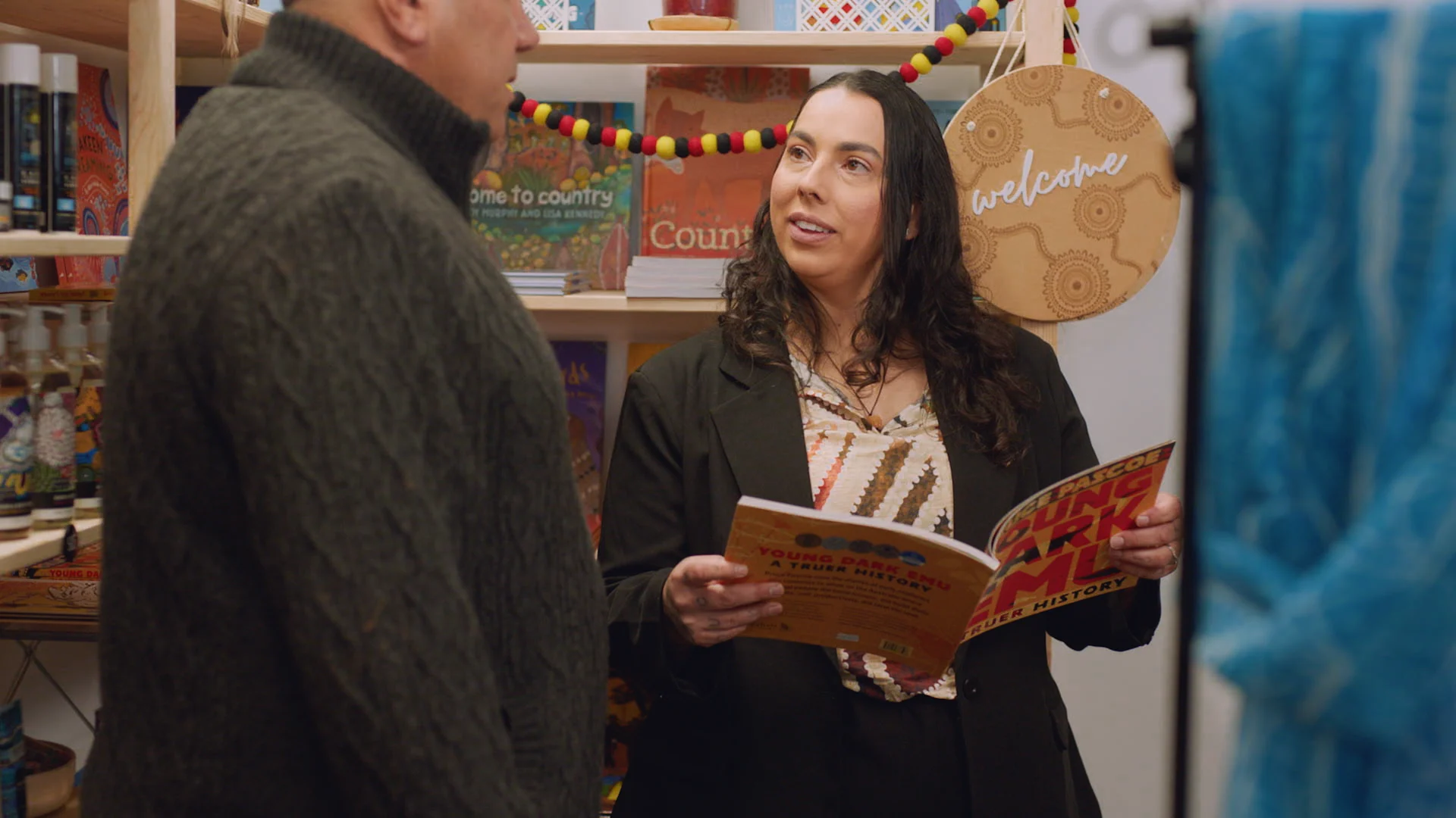 Two people conversing in a bookstore with Indigenous books and decorations, woman holding open book.