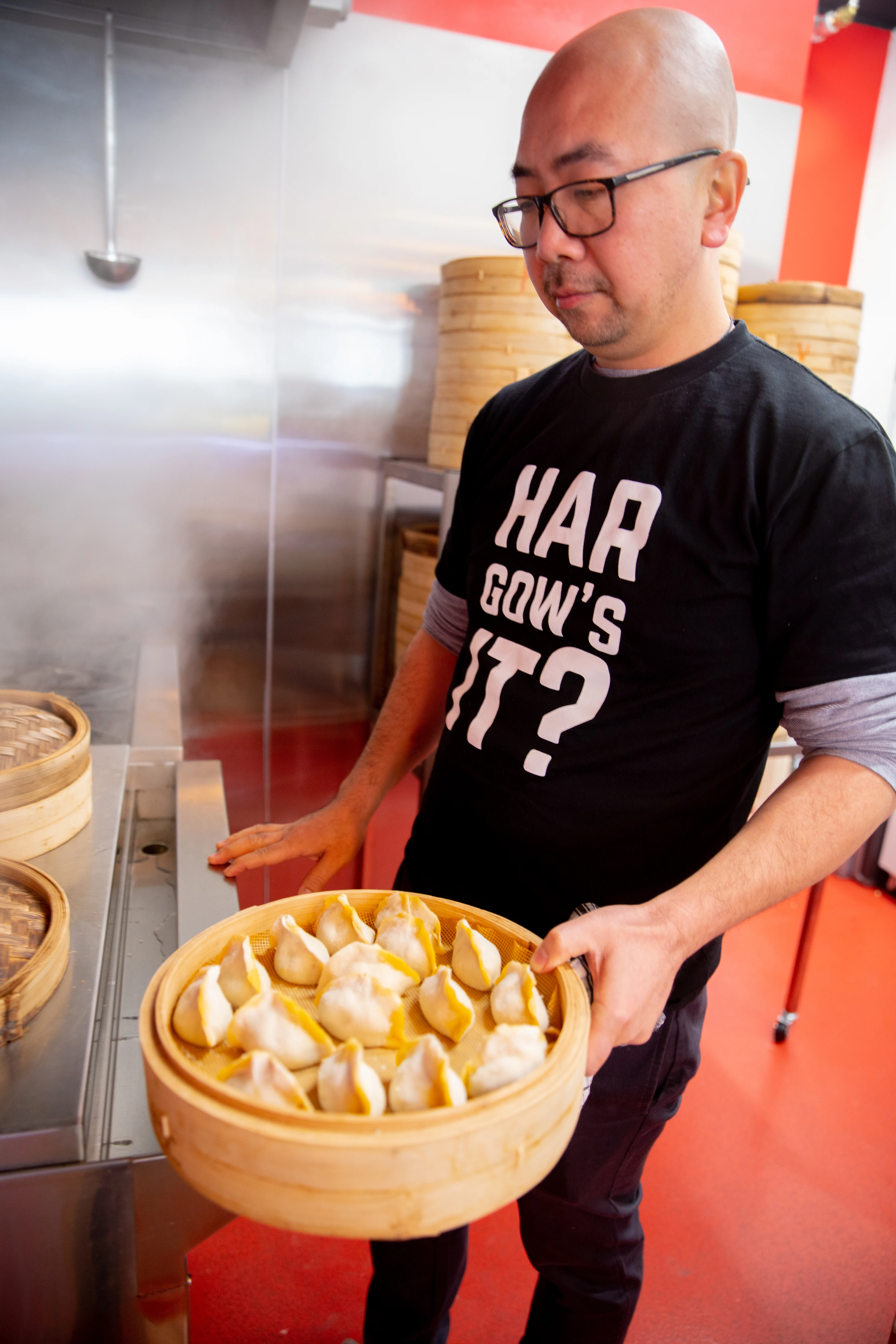 Gordon Kong stands in a commercial kitchen. He is holding a bamboo steamer filled with dumplings.