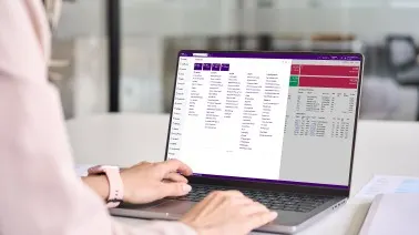 Person typing on laptop displaying data tables with purple and red interface sections in an office environment.