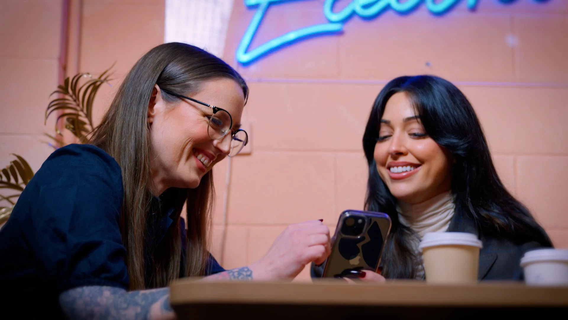 Two women smiling while looking at a smartphone together in a café with pink walls and blue neon sign.