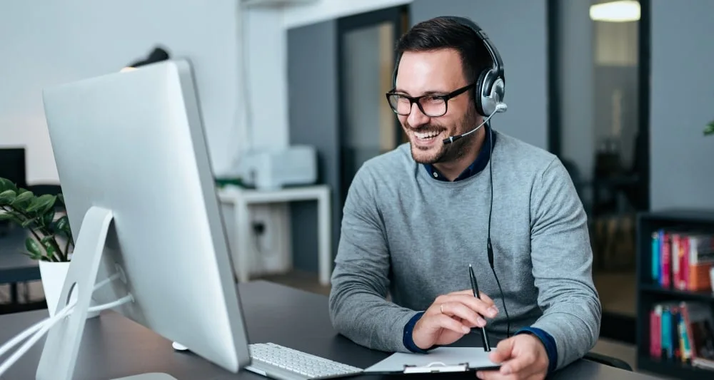 An employee wearing a headset is smiling. They are sitting at their desk and looking into their computer monitor, whilst holding a clipboard and pen.