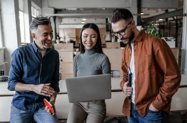 Three employees in an office are gathered together and smiling. The person in the middle holds a laptop, whilst all three look at the screen.