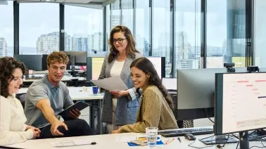 Diverse team of young professionals collaborating in a bright modern office with city skyline views through large windows.